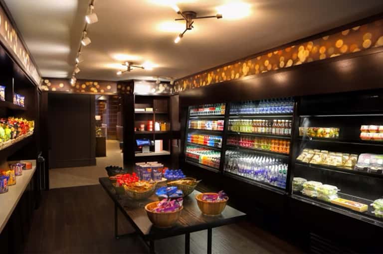 Modern convenience store interior with refrigerated beverage displays, product shelves, and central tasting table with snacks