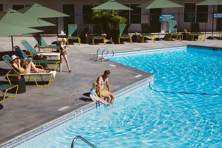 Resort pool deck with lounging guests, yellow umbrellas, green shade structures, and clear blue water