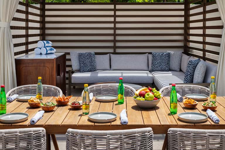 Upscale outdoor dining setup with striped privacy screen, gray seating, wooden table, and fresh fruit display