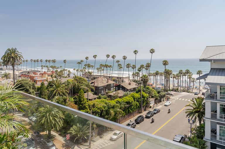 Coastal oceanfront view with palm trees, sandy beach, and street-level buildings overlooking the water