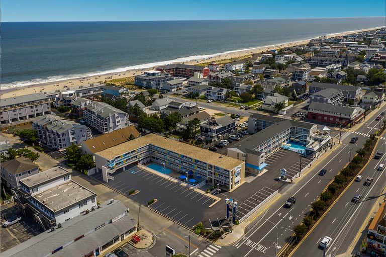 Aerial view of beachfront resort with pool, parking lot, and residential properties along sandy shore