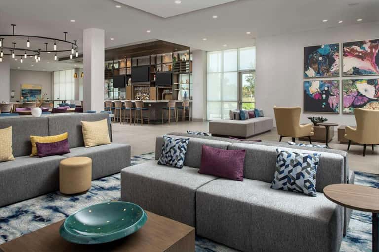 Modern hotel lobby with gray sectional sofas, colorful artwork, bar area, and large windows overlooking grounds