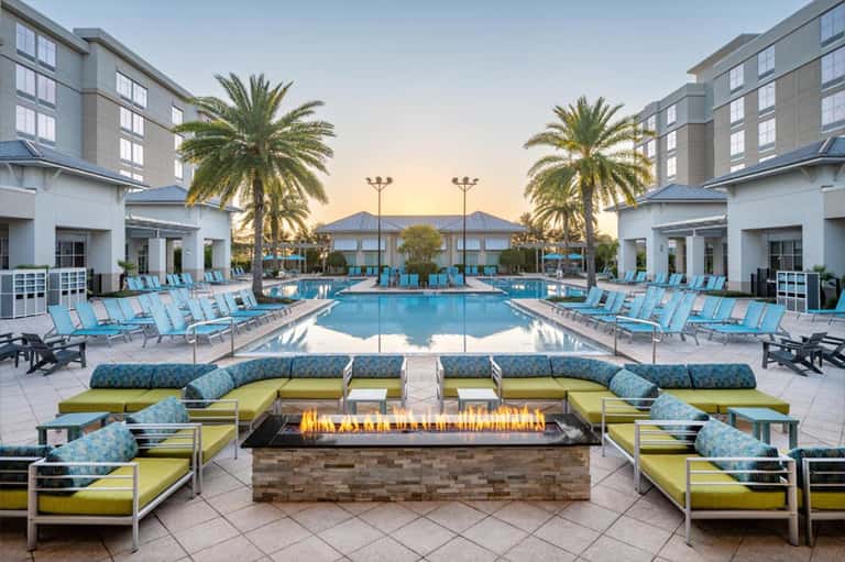 Resort pool at sunset with turquoise water, lounge chairs, firepit, and palm trees flanking modern buildings