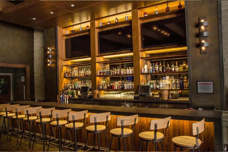Upscale bar with wooden counter, illuminated shelving displaying spirits, and comfortable seating
