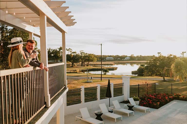 Couple on pergola balcony toasting with wine overlooking marshland and water with trees