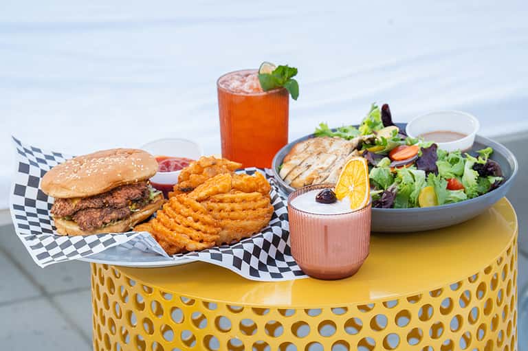 Gourmet burger, waffle fries, grilled chicken salad, and cocktails served on yellow table with ocean view