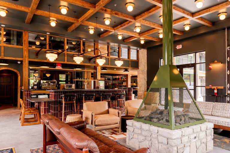 Rustic lodge interior with wooden beams, fireplace, leather seating, bar, and open gathering space