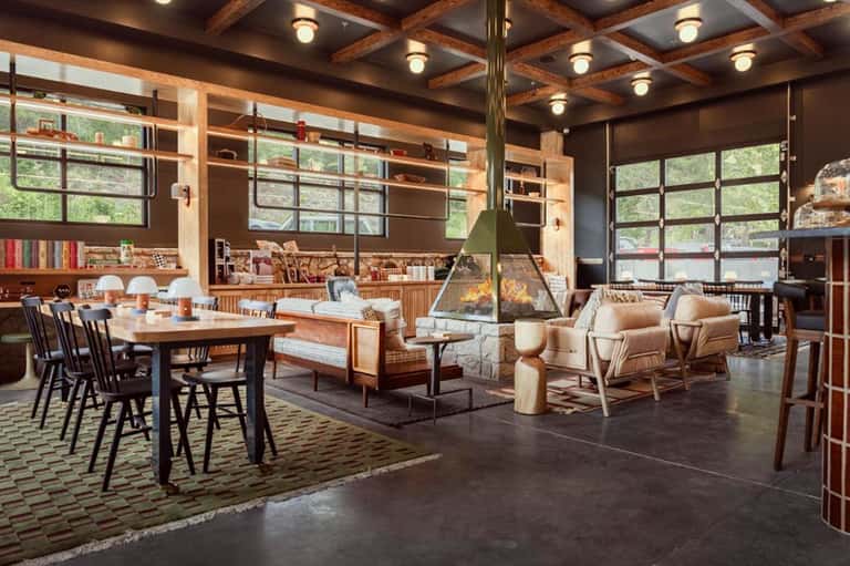 Rustic lodge interior with wooden beams, fireplace, dining area, and large windows overlooking forest