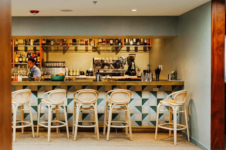 Contemporary coffee bar with white rattan bar stools, espresso machine, shelving, and geometric tile backsplash