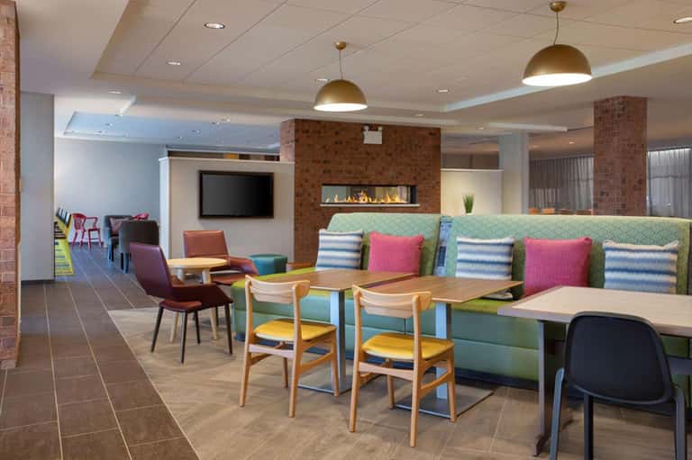 Modern hotel lounge with colorful seating, brick fireplace, pendant lights, and casual dining tables