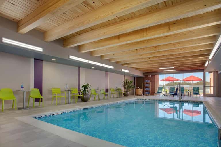 Indoor swimming pool with wooden beam ceiling, bright natural light, lime green chairs, and outdoor terrace view