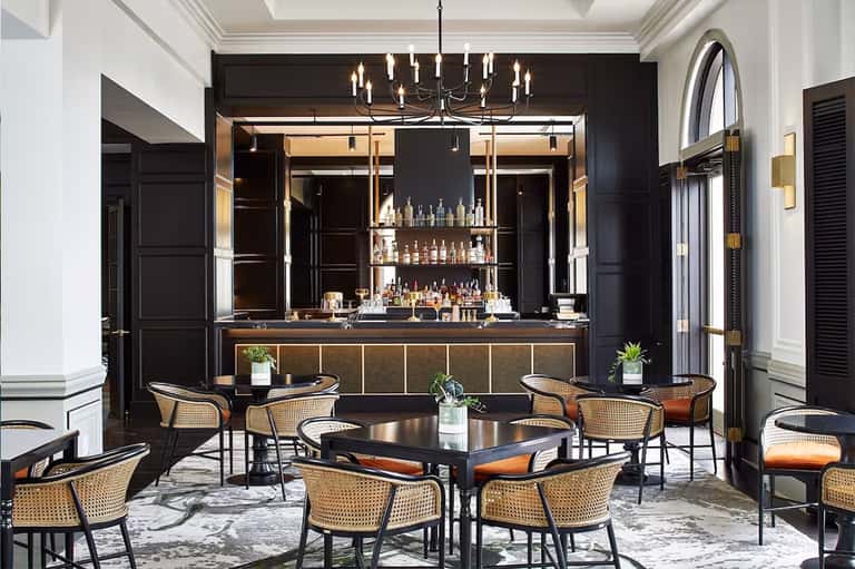 Upscale hotel bar lounge with black walls, illuminated bottle display, woven chairs, and modern pendant lighting