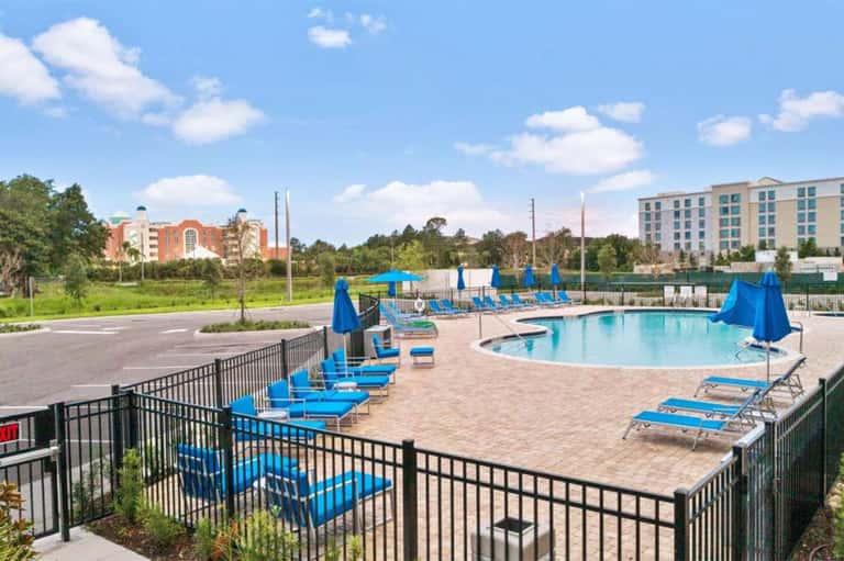 Outdoor resort pool with blue loungers and umbrellas, surrounded by landscaping and multi-story hotel building