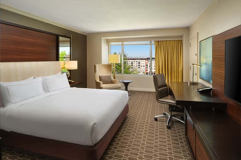 Hotel bedroom with queen bed, desk, chair, and city view from window