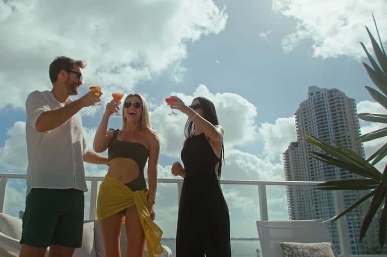 Three people toasting cocktails on a high-rise balcony overlooking city buildings and waterfront