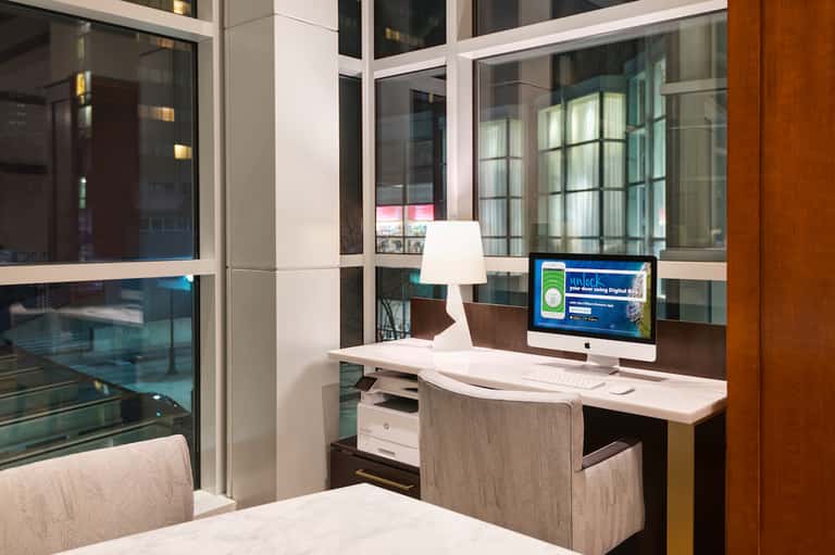 Hotel room desk with computer monitor and lamp, overlooking city skyline through floor-to-ceiling windows