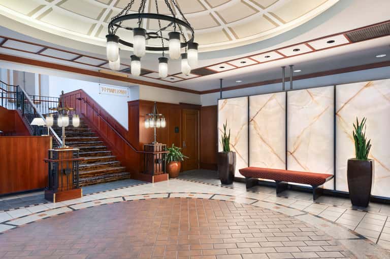 Grand hotel lobby with wooden staircase, marble floors, decorative plants, and pendant lighting
