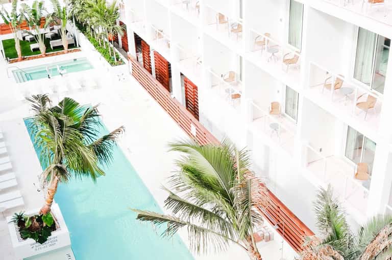 Overhead view of modern hotel courtyard with turquoise pool, palm trees, and white building