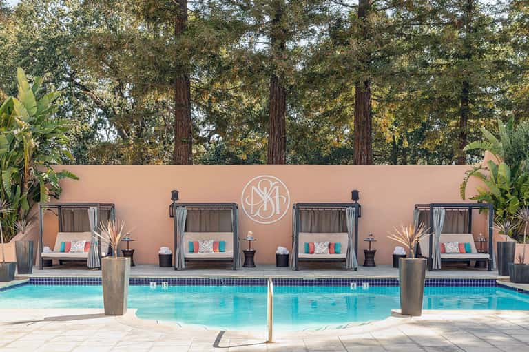 Luxury resort pool with cabanas and pink wall backdrop surrounded by tall trees at sunset