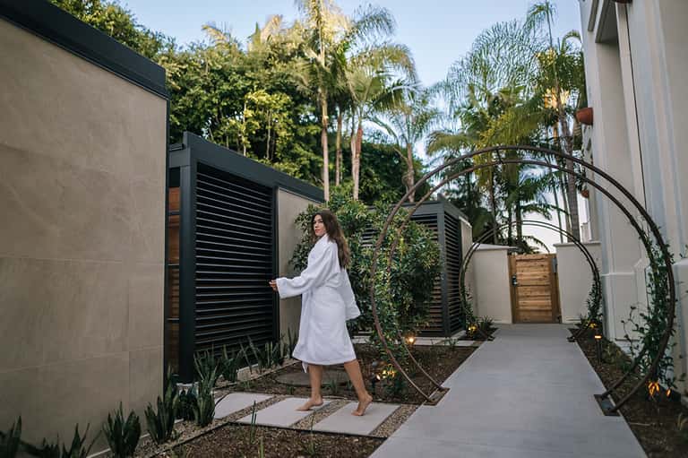 Woman in white robe walking through modern spa courtyard with black louvered gates