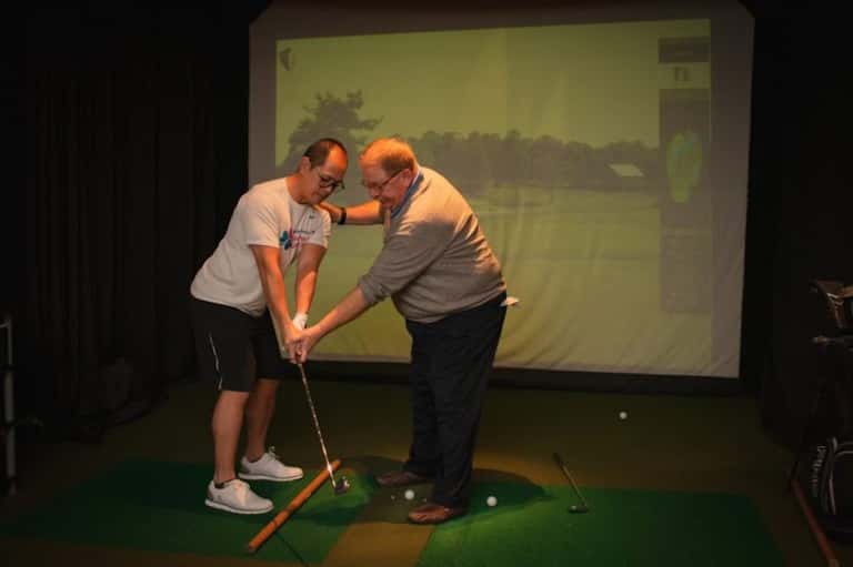 Two men playing indoor golf simulator with projected course display on screen