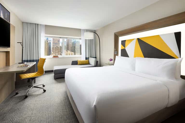 Modern hotel room with king bed, work desk, seating area, and city skyline view through large windows.