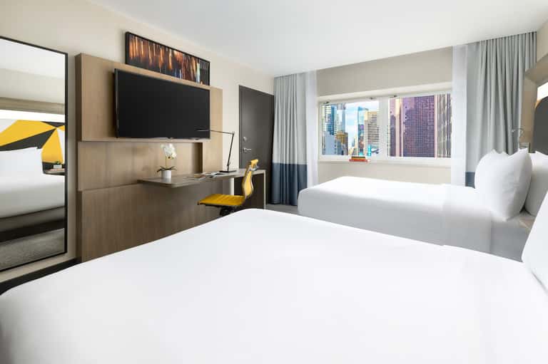 Contemporary hotel room with twin beds, flat-screen TV, wooden desk, and city skyline views through windows