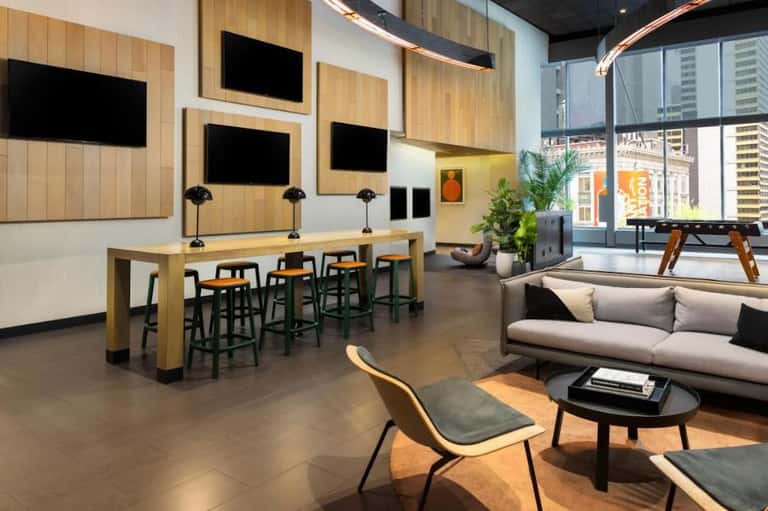 Contemporary hotel lounge with wooden accent wall, multiple TV screens, bar seating, sofa, and floor-to-ceiling windows