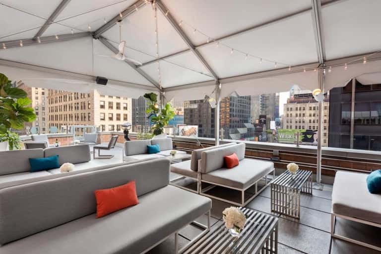 Modern rooftop lounge with gray seating, colorful pillows, city skyline views through glass walls, string lighting
