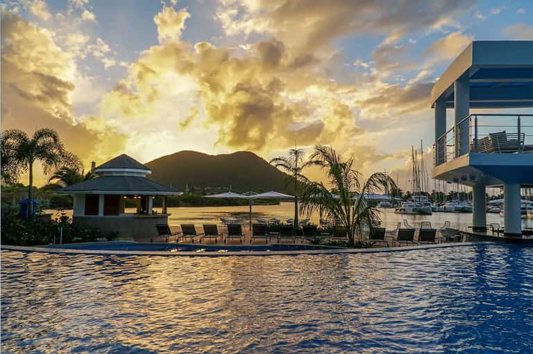 Waterfront resort with poolside cabana, mountain backdrop, palm trees, and moored sailboats at sunset