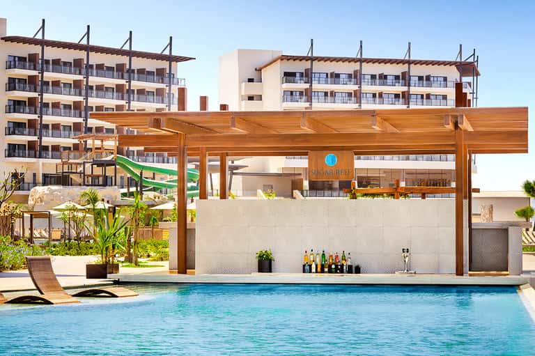 Resort pool with wooden pergola structure, swim-up bar, modern apartment buildings, and palm trees