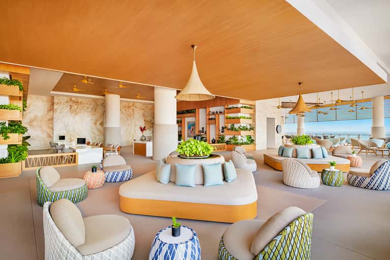 Modern resort lobby with orange ceiling, planted seating areas, pendant lights, and ocean views