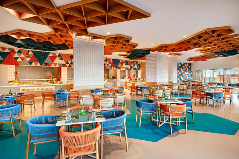 Bright dining room with geometric wooden ceiling panels, blue and orange furniture, and open kitchen