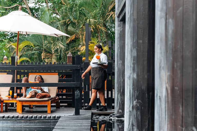 Beachfront restaurant patio with server, white umbrella, lounging guests, and tropical plants