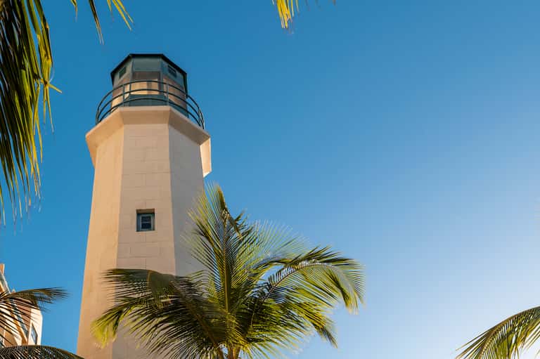 Tall white lighthouse tower with observation deck against clear blue sky and palm trees