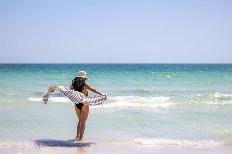 Woman in straw hat wading in turquoise ocean waves on sandy beach