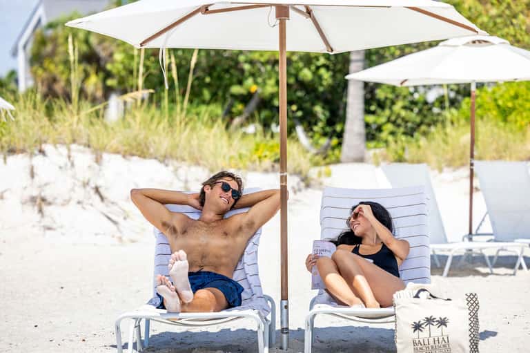Couple lounging under white umbrellas on a sandy beach with palm trees in background