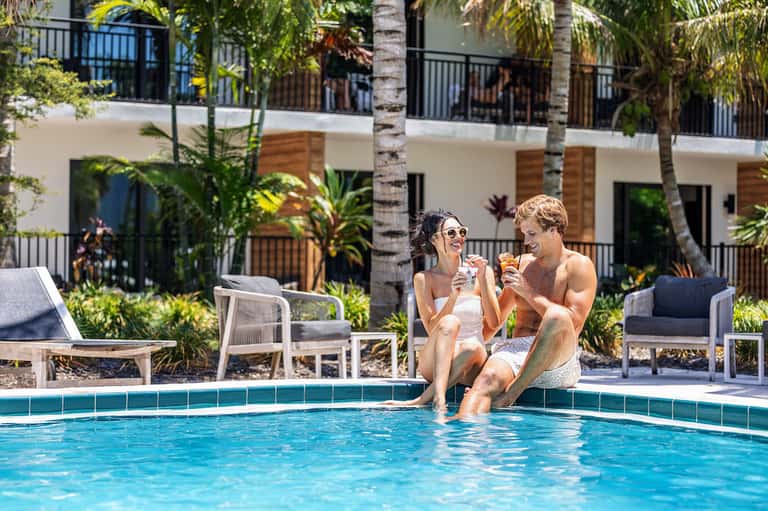 Two people sitting at pool edge with tropical resort building, palm trees, and loungers in background