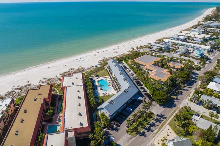 Aerial view of beachfront resort with pool, sandy beach, palm trees, and surrounding accommodations