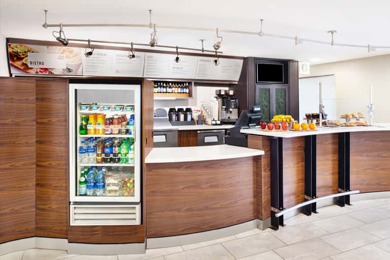 Modern hotel café counter with wood paneling, refrigerated beverage display, menu boards, and service area