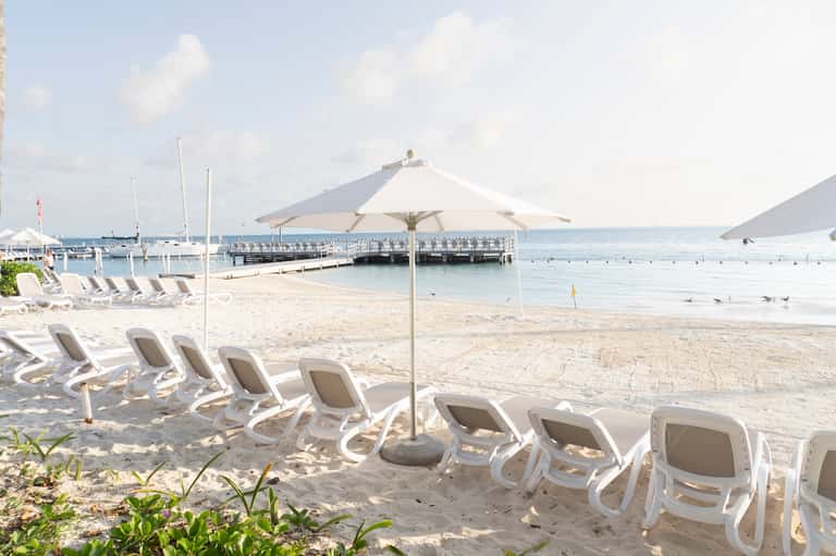 White lounge chairs with umbrellas on sandy beach with boats and pier in background