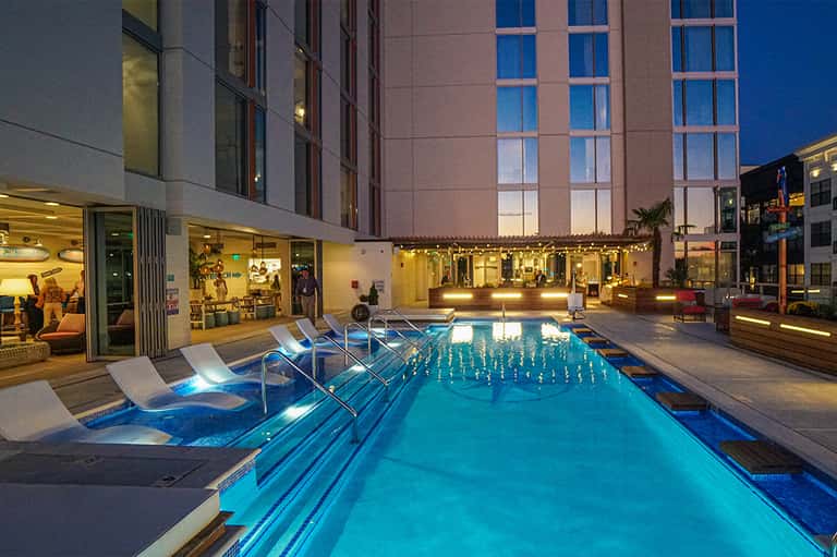 Illuminated indoor lap pool at dusk with lounge chairs, tall building facade, and evening lighting