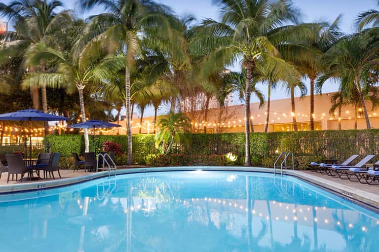 Resort swimming pool at dusk with palm trees, blue umbrellas, lounge chairs, and illuminated building backdrop.