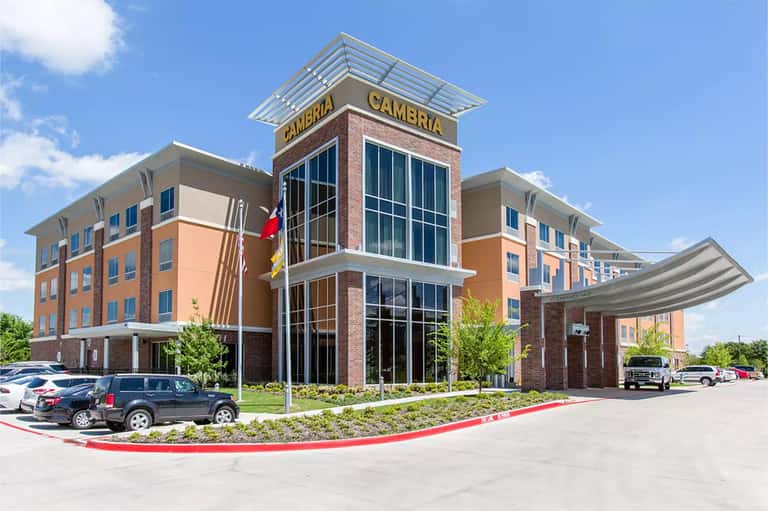 Modern hotel exterior with brick and orange facade, central tower marked Cambria, and parking lot