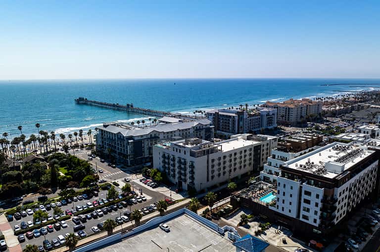 Aerial view of beachfront hotel complex with palm trees, pier, and sandy beach along California coast.