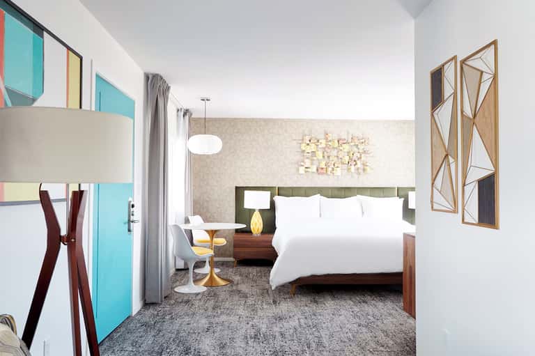 Modern hotel bedroom with green headboard, white bedding, contemporary furnishings, and colorful wall art