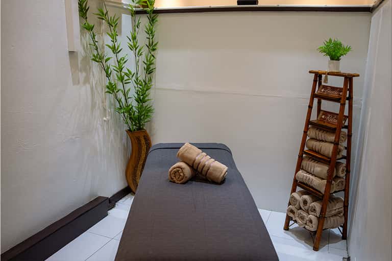 Spa massage room with gray massage table, rolled towels on wooden ladder, potted plants, and minimalist décor