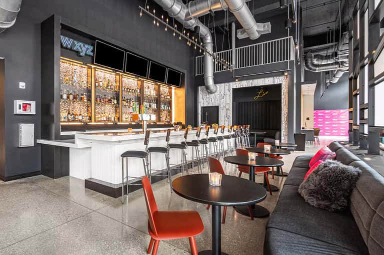 Contemporary bar and lounge with illuminated spirits display, white bar counter, red chairs, industrial ceiling