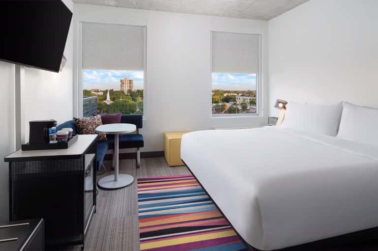 Modern hotel room with white beds, desk, city views through large windows