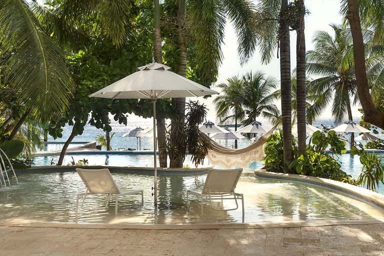 Tropical pool setting with white lounge chairs, umbrella, and hammock among palm trees with ocean view